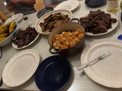 A family is having dinner at a table laden with food. The table is covered with a light-colored tablecloth. In the foreground, to the left, is a white oval platter filled with grilled corn on the cob. Next to it is another white oval platter with pieces of dark meat, possibly beef. A pot of cooked shrimp is in the center of the table, and a white oval platter with roasted potatoes is behind it. To the right is a white oval serving platter with a fork resting on it. A dark blue plate is in the lower right quadrant of the image. In the background, a person in a gray t-shirt is holding a phone, and another person is seated. A bottle of white wine stands on the table in the upper right corner. The scene appears to be indoors, likely in a home dining room.