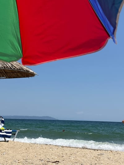 The image depicts a beach scene in Asprovalta, Greece. In the foreground, a beach umbrella with sections of green, red, and blue fabric is visible, casting a shadow. Below it, a straw umbrella is partially seen. The beach itself is sandy and covered with small pebbles. The sea is a deep green color, with white waves crashing gently on the shore. In the distance, a hazy line of mountains forms the horizon. A beach chair with striped towels is set up on the sand. A person can be seen swimming in the sea. The sky is clear and blue.