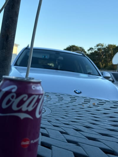 The image depicts a close-up of a purple can of Coca-Cola with a straw inserted, placed on a textured grey surface, possibly a table. In the background, a silver BMW car is visible, with a clear blue sky overhead and trees in the distance. The scene appears to be outdoors, with a light pole to the left. There is no discernible information within the image to suggest a specific location in Chichester, United Kingdom.