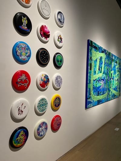 The image displays an art exhibition in Jinniu District, China. The foreground features a white wall adorned with a grid of decorative frisbees, each featuring a unique design. These designs range from cartoon characters and abstract patterns to an image of an alien and a dragon. The frisbees are arranged in rows, creating a visually interesting display. To the right of the frisbee display is a large, vibrant abstract painting with predominantly blue and green hues, accented by thick, neon green lines that appear to form characters. The painting is mounted on the wall and extends horizontally across a significant portion of the frame. The floor in the foreground is made of polished wooden planks. The lighting in the scene appears to be artificial, highlighting the artwork and creating subtle shadows on the wall. No people are visible in the image.