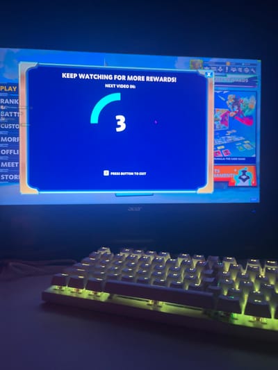The image depicts a gaming setup located in Donchevo, Bulgaria. An Acer brand computer monitor, partially visible, displays a video game interface. A prominent blue rectangular overlay occupies the central screen area, featuring white text "KEEP WATCHING FOR MORE REWARDS!" and "NEXT VIDEO IN:", accompanied by a light blue circular progress bar indicating a countdown of "3." Below the countdown, the instruction "Z PRESS BUTTON TO EXIT" is visible. Portions of the underlying game interface are visible on the left and right, including menu items such as "PLAY" and "RANK," and a section labeled "FREE REWARDS" with artwork for "BRAWLHALLA: THE CARD GAME." Positioned on a flat, dark surface directly in front of the monitor is a white, compact mechanical keyboard. Its keys are illuminated with a yellowish-green backlight. The ambient lighting in the environment is low, with the monitor and keyboard providing the primary illumination. No individuals or active interactions are discernible within the frame.