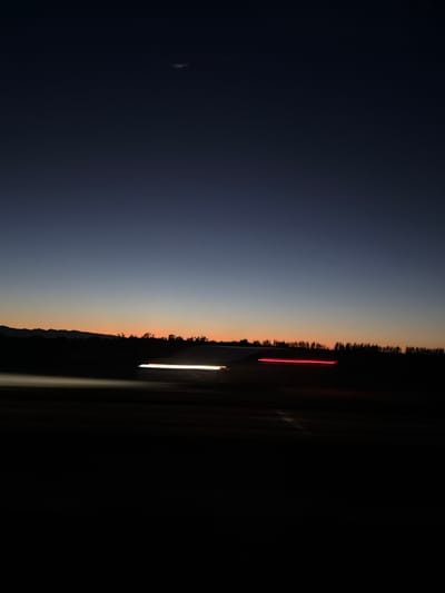 At dusk, a dark landscape is illuminated by streaks of light from passing vehicles. The sky transitions from deep blue at the top to a pale orange near the horizon, where a silhouetted mountain range is visible. The foreground is dominated by darkness, with the light trails indicating movement on a road or highway. The location is identified as Dixon, United States.