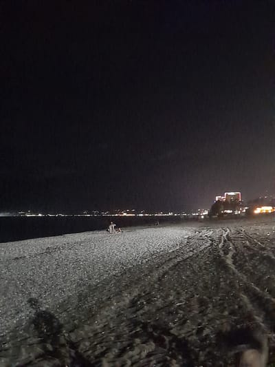 The image depicts a nighttime scene on a beach in Psakhara, Abkhazia. The sky is dark and devoid of stars. A dark body of water is visible in the background, with a distant shoreline illuminated by a strip of lights, suggesting a populated area or city. A prominent building with illuminated windows, appearing to be a hotel or large structure, is situated on the right side of the distant shoreline.

The beach itself is covered in pebbles or small stones, with tire tracks visible in the sand towards the foreground and right side of the image. In the middle ground of the beach, several individuals are present. Two figures appear to be standing, and a third person is seated or lying down. They are positioned relatively close to each other. The overall atmosphere is dark and quiet, with artificial lights providing the primary illumination.