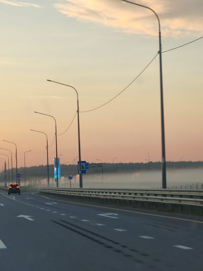 The image shows a highway at dawn in Bolshiye Dvory, Russia. A single car is driving on the road, and there are several streetlights lining the highway. The sky is a pale orange and yellow, with some clouds. There is fog in the distance, obscuring the treeline. Road markings include white arrows indicating lane direction and dashed white lines separating lanes. A guardrail runs along the side of the highway. Several road signs are visible, including a blue sign with white text and a blue sign with a white tree symbol. The overall scene suggests a quiet, early morning on a major road.