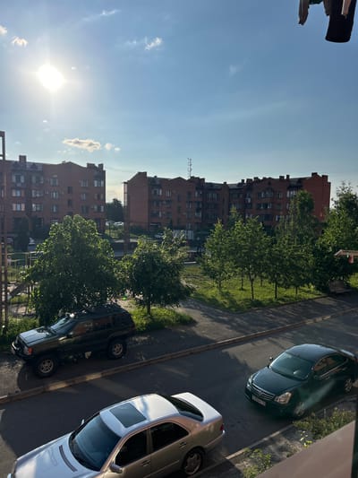 The image shows a view from a height, overlooking a street and residential buildings. Two multi-story brick buildings with many windows are prominent, likely apartment complexes. A street runs in front of them, with two cars parked on it: a dark-colored SUV with a roof rack in the foreground on the left, and a dark sedan parked further down the street on the right. Green trees and grass are visible between the buildings and the street, with some areas appearing to be a playground or park. The sky is blue with some scattered clouds and a bright sun. Clothing is seen hanging to dry in the upper right corner of the image. This scene is located in the city of Beslan, Russia.