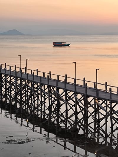 This media file captures a serene waterscape at dawn or dusk, likely in Labuan Bajo, Indonesia. The scene is dominated by a long, wooden pier stretching from the foreground into the calm water. The pier, constructed with sturdy wooden pilings and a railing, is sparsely illuminated by a few lampposts. Its reflection is vividly mirrored in the water below, creating a striking pattern of light and shadow.
In the distance, a small, colorful boat with a canopy is gently floating on the water. Beyond the boat, a hazy mountain range is visible on the horizon, softened by the atmospheric conditions. The sky displays a gradient of soft orange and pink hues, indicative of either sunrise or sunset.
The overall mood is peaceful and tranquil. There are no people visible in the frame, emphasizing the quiet solitude of the moment. The water appears still, reflecting the muted tones of the sky and the pier. The weather seems calm, with no indication of wind or rough seas.
Notable details include the rustic construction of the pier, the distant, ethereal mountains, and the solitary boat adding a touch of color to the otherwise muted palette. The time of day is clearly suggested by the warm, soft lighting and the gradient of colors in the sky. The location context of Labuan Bajo, Indonesia, is a plausible interpretation given the tropical setting and the type of boat. No visible text is present in the image.