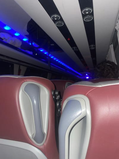 The interior of a passenger vehicle, likely a bus or plane, is shown. Rows of cushioned seats in maroon and gray hues are tightly aligned. A strip of vibrant blue LED lights illuminates the narrow aisle. The wall beside the seats is white with horizontal black stripes, punctuated by small, circular air vents. A glimpse of curly hair appears between the wall and the seats.