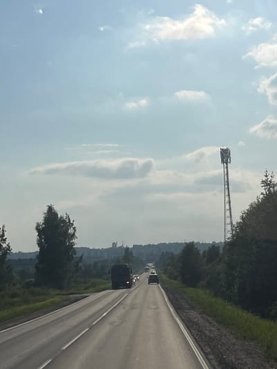The image, captured from a moving vehicle on a highway in Romanovka, Russia, depicts a scene on a cloudy day. The primary subject is a cell tower, a tall, lattice-like structure with antennas at its apex, dominating the left side of the frame. The tower stands against a pale blue sky filled with soft, wispy clouds. To the right of the tower, a dense line of trees forms a natural border along the highway.
The highway itself stretches into the distance, with several vehicles visible on it. The road appears to be a multi-lane thoroughfare with white lane markings. The right side of the highway is bordered by a grassy embankment with patches of rough terrain. The time of day appears to be daytime, given the brightness of the sky and the visibility of the landscape. The weather seems to be overcast, as indicated by the diffused light and the presence of clouds.
The overall mood is one of travel and infrastructure in a rural or semi-rural setting. The image conveys a sense of movement and the passage of time, as suggested by the perspective from a moving vehicle and the highway leading into the distance. There are no visible people, text, or specific emotional cues present in the image.