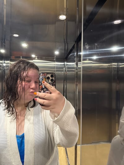 The image, captured in Tulovo, Bulgaria, depicts a female subject taking a mirror selfie within what appears to be an elevator.
The subject is a woman with dark, wet, and visibly disheveled hair, which hangs loosely around her face and shoulders. Her skin appears somewhat flushed. She is making a puckering or "kissing" expression with her lips and her eyes are directed towards the smartphone screen. She is wearing a white, waffle-knit bathrobe, partially open to reveal a blue garment underneath, possibly a swimsuit top or tank top. Her left hand is prominently visible, holding a dark-colored smartphone, likely an iPhone model due to the distinct triple-lens camera array. The phone is encased in a protective cover featuring a collage of multiple small photographic images, predominantly depicting various faces and individuals. The word "WOMBLOOK" is discernibly printed in reverse on the phone case, indicating a reflection. Her visible fingernails are painted; the thumbnail is a solid bright orange, while the index finger displays a red and white patterned design.
The environment consists of highly reflective, brushed metallic surfaces forming the walls and ceiling, consistent with an elevator interior. Several circular, recessed downlights are visible on the ceiling, emitting bright light. The metallic surfaces show various reflections, including horizontal light streaks and apparent smudges or marks. A vertical dark seam or panel division is visible on the right side of the reflective surface. The lower portion of the reflection on the right wall indicates a muted, light brown or beige color, potentially a floor or lower wall panel outside the direct frame.