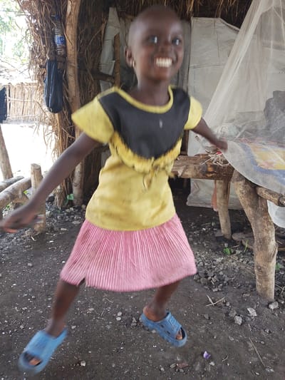 The image shows a young girl in Otulu, Nigeria, outdoors. She is wearing a yellow and black top and a pink pleated skirt. She is also wearing blue sandals. The girl is smiling and appears to be mid-movement, possibly dancing or playing. Behind her, a rustic structure is visible, constructed from branches and thatch, with a water bottle and a black bag hanging from one of the supports. A white mosquito net is draped over a wooden structure to the right, suggesting the presence of a sleeping area or shelter. The ground is dirt with small stones. The lighting indicates it is daytime.