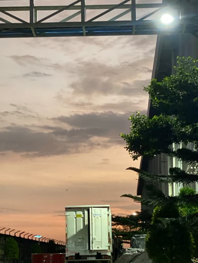 The image depicts a twilight scene in Gambut, Indonesia, characterized by a soft, peachy sky with scattered grey clouds. In the foreground, the right side of the frame is dominated by the dark silhouette of a building and lush green foliage, illuminated by a bright overhead light. To the left, a white cargo trailer with its door slightly ajar is visible, positioned behind a chain-link fence with a serrated top. A red cylindrical object is partially visible in the lower left corner. The overall impression is one of an industrial or commercial area transitioning into evening.