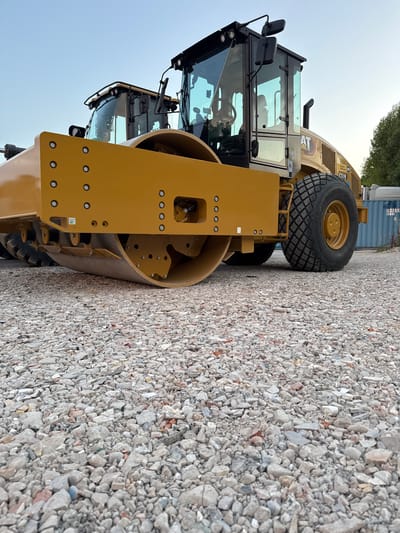 Two yellow Caterpillar CS19 vibratory soil compactors are parked on a gravel lot. The compactor in the foreground is positioned at an angle, showing its large roller drum and rear tire. The second compactor is partially visible behind it. The sky is clear and blue. The scene is located in Grenay, France.