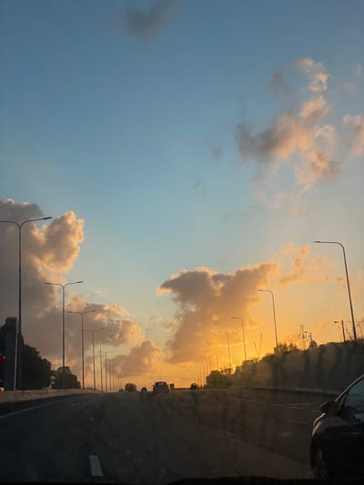 The image depicts a view from a vehicle moving along a road. The sky is visible, transitioning from a light blue to orange-yellow towards the horizon. Clouds are present, illuminated by the sunset, appearing white and greyish. A line of streetlights flanks the road. On the right side of the frame, a partial view of the road and other vehicles can be seen. Based on the information provided, this is located in Hod HaSharon, Israel.