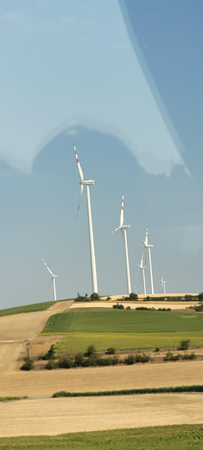 A landscape photograph, taken from a slightly elevated perspective, captures a field of wind turbines against a clear blue sky with some wispy clouds. The wind turbines, predominantly white with red accents at the tips of their blades, are spread across rolling hills. In the foreground, fields of varying textures and colors suggest agricultural activity, with patches of brown earth and green crops. A dirt road or path winds its way up a grassy hill on the left side of the image. The scene appears to be outdoors in a rural or semi-rural area. The metadata indicates the image was taken in Hobersdorf, Austria.