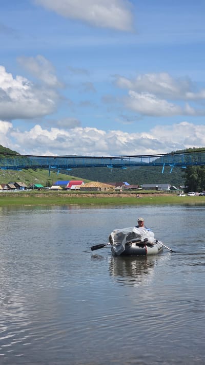 In the foreground, a person wearing a camouflage cap and a light-colored shirt is paddling a gray inflatable boat across a body of water. The boat is loaded with items covered by a translucent plastic sheet. The water's surface is rippled, reflecting the sky and clouds. In the middle ground, a village with houses of various colors, including red, blue, and green roofs, is situated on a grassy bank. A structure under construction with exposed wooden beams is also visible. A blue suspension bridge spans across a hill covered in greenery in the background, beyond which are forested mountains. The sky is a bright blue with scattered white clouds. The scene is located in the village of Tash-Asty, Russia.