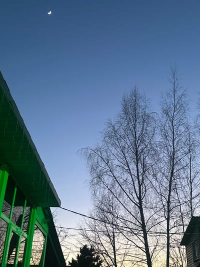 The image displays an outdoor scene at either dawn or dusk in 3-Y Uchastok, Russia. The sky transitions from a deep clear blue at the zenith to a pale yellow-orange near the horizon. A thin crescent moon is positioned in the upper left quadrant of the sky. In the lower left, a portion of a building with a vibrant green exterior, sloped roof, and multiple windowpanes is visible. Small, white, icicle-shaped decorative lights hang from its eaves. A single thin, dark cable extends horizontally from the corner of this green building towards the right. To the right, several tall, bare deciduous trees, possibly birches, are silhouetted against the gradient sky. A darker evergreen tree line is discernible in the lower background. A fragment of a second, lighter-colored wooden building is present in the lower right corner. No people, specific actions, or interactions are visible.