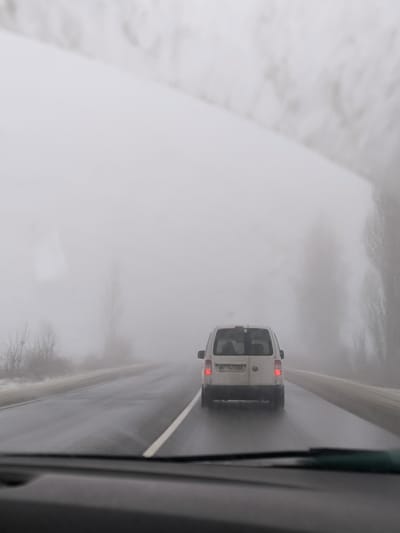 The image depicts a foggy winter or late autumn road scene in Khotyn, Chernivtsi Oblast, Ukraine, viewed from the interior of a vehicle. A light-colored Volkswagen Caddy-type utility van, bearing Ukrainian license plate "BT 9633 BO" (indicating Chernivtsi Oblast), is being followed on a wet, two-lane asphalt road. Both of the van's rear brake lights are illuminated. Heavy fog significantly reduces visibility, obscuring distant features. Bare trees and sparse vegetation are faintly discernible along the roadside, with traces of snow or frost on the ground. The foreground shows the top portion of a vehicle's dashboard and the lower edge of its windshield, through which the scene is observed.