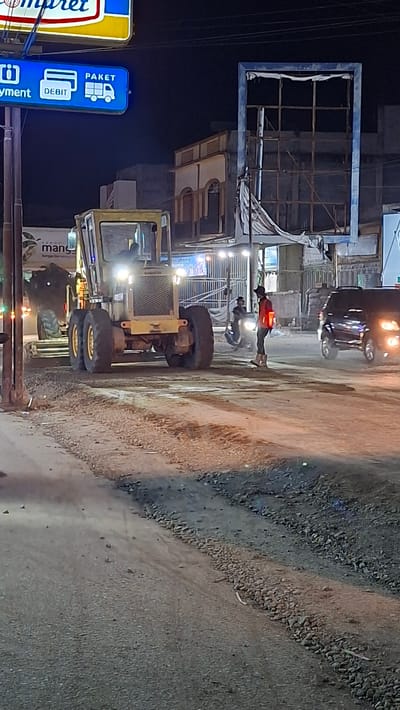 A nighttime scene in Blangpeuria, Indonesia, depicts a street with several vehicles and people. The image is vertically oriented. In the foreground, a yellow backhoe loader is parked, its large tires prominent. To the left, a blue and yellow sign for "Indomaret" is visible, featuring the text "PAKET" and "DEBIT" along with icons for a delivery truck and credit cards. Further back, a dark-colored car with illuminated headlights is present. Several individuals are scattered throughout the scene. One person, wearing a red shirt, stands near the car. Another person is seen riding a motorcycle. The environment appears to be a street with a rough, gravelly surface. The lighting suggests it is nighttime, with artificial lights illuminating the area. The overall atmosphere is that of a typical street scene in a semi-urban or rural Indonesian setting at night.