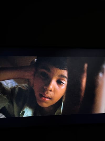 A child rests their head on an arm, looking up with a slightly tilted gaze. Their features are soft, with dark hair and eyes, and slightly parted lips. Light reflects off their skin, creating highlights on their face. They are dressed in a light-colored shirt or garment. The background is dark and blurry, creating a sense of intimacy and focus on the child. Part of a larger arm is visible above the child. The lighting seems dim and warm.