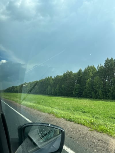 The image captures a view from a moving vehicle looking out a side window. In the foreground, the side mirror of the car is visible, reflecting a gray interior. To the left, a portion of the car's door frame and the asphalt road with a white lane marker are shown. Beyond the road, a vast, vibrant green field stretches towards a dense line of dark green pine trees. The sky above is a mix of bright blue with thin white contrails and dramatic, dark storm clouds gathering on the left side, casting a shadow over a portion of the field. The scene depicts a rural landscape outside of Irnykshe, Russia, under a dynamic sky.