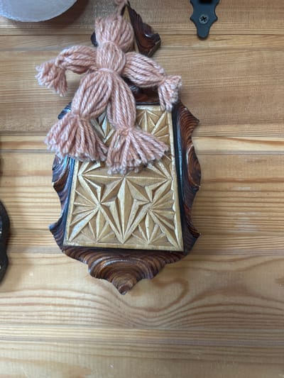 A close-up, overhead shot shows a wooden plaque mounted on a wooden wall. The plaque features a carved starburst design within a darker wooden frame that has a scalloped edge. A pink yarn doll, made from multiple strands tied together, is attached to the top of the plaque. The doll has a circular head, and two outstretched arms with tassel-like hands. Below the arms, two tassel-like legs are attached. The wooden wall is constructed of horizontal planks with visible grain. A black metal bracket with a screw is visible above the plaque, attached to the wall. The scene is likely an interior decoration, possibly in a home or workshop. There is no information available regarding the specific location in Mar'ino, Russia.