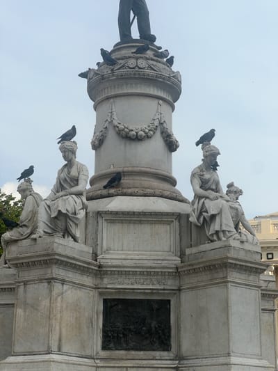 This image shows the Monument to the Heroes of 1811 in San Salvador, El Salvador, seen from a low angle. The monument features a tall column topped with a statue of a figure. The base of the column is adorned with sculpted garlands and is occupied by numerous black birds, likely pigeons or crows. At the base of the monument are marble statues of draped female figures in classical style. The monument itself is made of light-colored stone or marble. In the background, the sky is overcast. A building partially visible in the upper right corner, is painted white. The monument is likely located in a public square or plaza within the city.