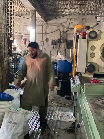 The image depicts an industrial workshop located in Daman, Afghanistan. In the foreground, a bearded man, dressed in a two-tone light brown and olive-green traditional tunic over trousers, is captured mid-stride, moving towards the left. His head is slightly blurred, indicating motion.
The environment is a utilitarian industrial space. The walls are unpainted concrete or blockwork, exhibiting visible wear, grime, and haphazardly run electrical wiring, including several wall-mounted electrical control boxes. The ceiling features exposed corrugated material or rebar structure. The concrete floor is noticeably wet in sections and partially covered with corrugated metal sheets.
On the right side of the frame, a large industrial machine, possibly an injection molding or press unit, dominates. It features a prominent yellow structural element, grey control panels, and a red indicator light at its top. A control panel with multiple buttons and switches is integrated into the machine's side. A coil of red, possibly plastic, material rests on a flat surface of this machine.
In the left foreground, a heavy-duty chain hoist with a hook hangs vertically near a concrete pillar. Adjacent to it are stacked white plastic buckets and several large white plastic bags, some containing granular material and one partially labeled "P E". Further back on the left, a tangle of pipes, hoses, and electrical conduits is visible, alongside blue cylindrical drums or barrels. A red wall-mounted fire extinguisher is visible on the rear wall. Artificial lighting, including a bright bare bulb, illuminates the scene.