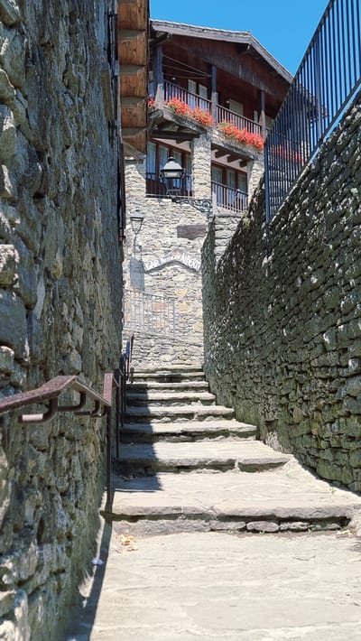 The image depicts a narrow, cobblestone alleyway in the town of Rupit i Pruit, Spain. Stone walls flank both sides of the alley, with a steep staircase made of large, flat stones leading upwards. To the left, a weathered metal handrail runs along the staircase. On the right, a portion of a building is visible, constructed with stone and wood. This building features balconies adorned with vibrant red flowers and a dark wooden facade. A sign is mounted on the stone wall, partially obscured, with text visible that appears to read "BAR RESTAURANT ALBER..." indicating it is a dining establishment. The sky above is a clear, bright blue, suggesting a sunny day. The overall scene conveys a sense of rustic charm characteristic of historic European villages.