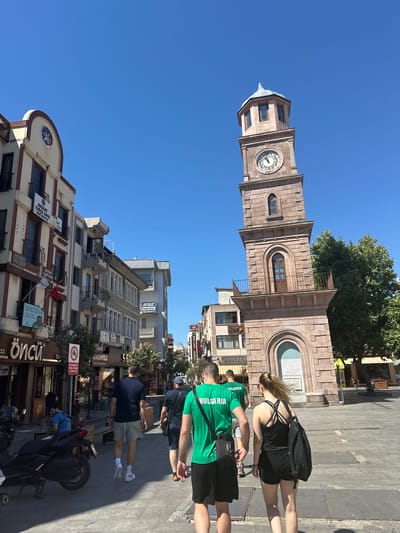 This media file captures a street scene in Çanakkale Merkez, Turkey, on a bright, sunny day, indicated by the clear blue sky and strong shadows. The primary focus is a prominent clock tower, a classic stone structure with arched windows and a visible clock face. In the foreground, several people are walking or standing on a paved walkway. One person wears a green t-shirt with "BULGARIA" printed on it, suggesting a connection to the country, possibly tourists or participants in an event. Another person is in a black tank top and shorts, carrying a backpack. Various buildings line the street, featuring storefronts with signage. Some of the visible text includes "AVUKAT" (lawyer) in Turkish, along with phone numbers and names, suggesting a commercial area. Other signs mention "PASTANESI" (pastry shop) and a motorcycle parking restriction. The overall atmosphere appears to be lively and typical of a town center with people going about their day.