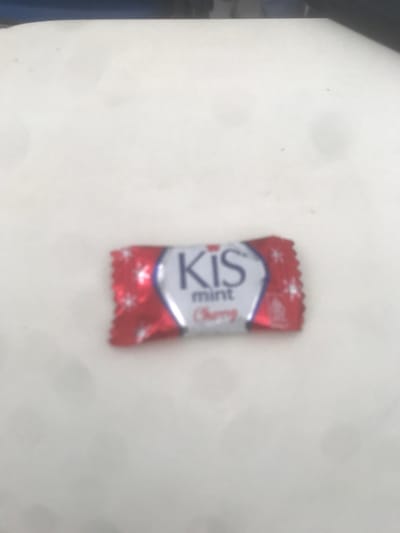 The image displays a single KIS mint Cherry candy, individually wrapped in a red, white, and silver foil. The wrapper features "KIS" prominently in blue lettering, with "mint" and "Cherry" (in red script) positioned below. Small white, four-pointed star or snowflake-like designs adorn the red portions of the wrapper. The candy rests centrally on a flat, uniformly light-colored surface, likely white or off-white. No other objects, individuals, or discernible actions are present in the frame. The scene is static. The location is Muka, Indonesia.