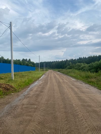An unpaved road extends into the distance, bordered by lush green grass and trees. A vibrant blue metal fence runs along the left side of the road, with utility poles and wires overhead. The sky is filled with dramatic, cloudy formations. The road's surface shows clear tire tracks, indicating recent vehicle traffic. The scene appears to be on the outskirts of Заволжский, Russia.