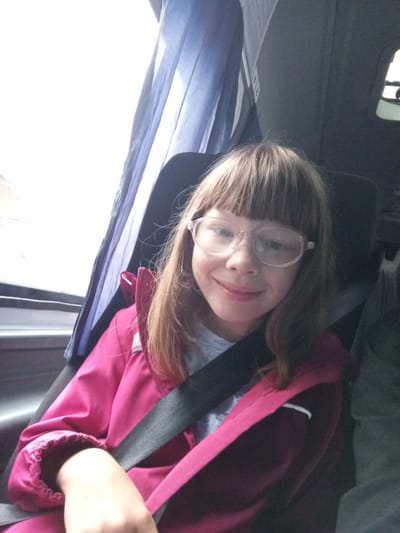 In Verkhneyarkeyevo, Russia, a young girl with blonde hair and bangs, wearing glasses and a pink jacket, sits in a bus seat. She is wearing a seatbelt and looking at the camera with a slight smile. The bus interior is visible, with blue curtains to the left and a dark gray seat behind her. The lighting suggests it is daytime.
