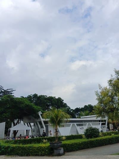 This image depicts a public leisure area in thành phố Biên Hòa, Vietnam, under an overcast sky. The primary subject is a modern, white, angular building featuring multiple "WC" (Water Closet) signs, indicating its function as public restrooms. The building has a distinctive geometric design with sloped walls and horizontal paneling.
In the foreground, a low, manicured green hedge defines a landscaped section containing a potted palm tree on a dark pedestal. A paved path or plaza surface is visible in the immediate lower foreground.
Several people are present. On the left side of the scene, a white canopy tent is erected, under which multiple individuals (approximately 3-4) are visible, seated or standing, alongside several parked bicycles or scooters. Near the left section of the main building, two individuals are standing. Further right, near another "WC" sign, two more individuals are standing, one in a light shirt and dark pants appears to be gesturing. In total, approximately 7-10 individuals are observed engaging in passive activities such as congregating or resting.
The background and upper sections of the image are filled with dense, dark green foliage from numerous trees, suggesting a park-like or heavily vegetated environment. The sky is largely covered with white and grey clouds, with small patches of lighter sky visible, indicating a cloudy day.