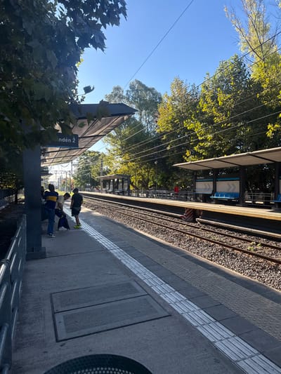 The image displays a train station platform on a sunny day, identified as Don Bosco station in Partido de Quilmes, Argentina. The foreground features a concrete platform with white tactile paving strips along its edge, beneath a modern, angled canopy supported by a grey column labeled "Andén 2". A silver metal railing extends along the left. Three individuals are on this platform: one adult wearing a blue and yellow striped shirt and blue jeans, leaning against the railing; one younger male in a light shirt and dark pants bending over near a blue bag; and another younger male in a dark shirt, dark shorts, and yellow-green sneakers, standing. A silver, perforated trash can is partially visible in the bottom center. Two parallel train tracks, covered in ballast, separate this platform from another identical concrete platform on the right midground. This second platform features a smaller canopy structure with blue benches and a sign indicating "Don Bosco". Abundant green foliage from numerous trees lines both sides of the tracks, and some branches overhang the foreground platform. Multiple overhead wires span the clear blue sky.