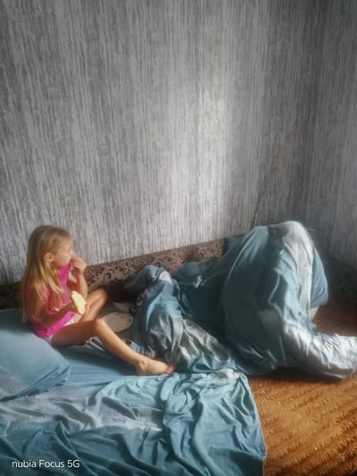 In the image, a young girl with blonde hair, wearing a pink top and white shorts, sits on a bed and eats a piece of food. She is looking to her right. Partially covered by a blue blanket to her right is another person, whose face is not visible. The bed has a patterned headboard and is covered with blue sheets. Behind the bed, the wall is covered with a textured wallpaper that appears to have a grey and white vertical stripe pattern. The room is dimly lit, suggesting it might be evening or a cloudy day. The text "nubia Focus 5G" is visible in the bottom left corner of the image. The setting appears to be a bedroom in Sestrynivka, Ukraine.
