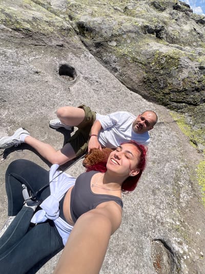 A high-angle, self-taken photograph captures a man, a woman, and a small dog posing on a large, weathered rock surface under bright daylight in Lenovo, Bulgaria. The woman, on the right and holding the camera, has vibrant red hair and is wearing a dark grey ribbed sports bra, black leggings, and a white long-sleeve shirt tied around her waist. She smiles broadly with closed eyes, showing a happy expression. She wears a silver necklace with a small pendant and stud earrings.

To her left, a man with a bald head and a short beard wears a white long-sleeve shirt with the text "OFF" visible on the chest, olive green shorts, and white sneakers. He has a red beaded bracelet on his wrist and looks towards the camera with a neutral to slightly smiling expression. He holds a small, brown, curly-haired dog, possibly a poodle, which is attached to a leash.

The setting is an outdoor, rocky landscape, likely a mountain or large rock formation. The massive grey rock surface where they are seated and surrounded by is covered with patches of greenish-yellow lichen. Two distinct, almost circular holes or depressions are visible in the rock, one near the man's left leg and another larger one to the right, which contains some natural debris. A faint "X" mark is also visible carved into a rock surface in the background. The clear sky is visible in the upper left corner of the scene, indicating sunny weather. The overall mood is relaxed and joyful.