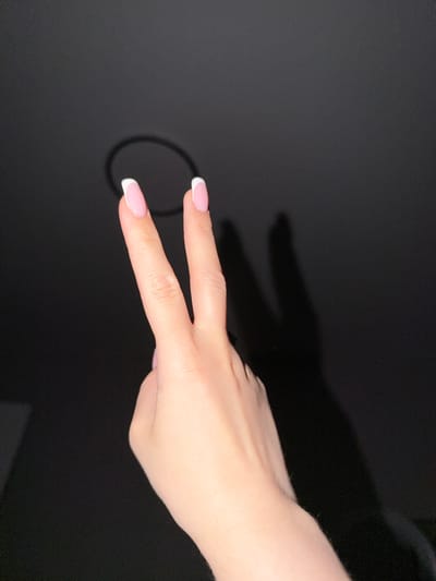 A left hand with French manicured nails is held up against a dark background, forming a peace sign with the index and middle fingers extended. A circular shadow, likely cast by a ring or other object, is visible behind the hand. The scene is indoors, and no specific location in Razumnoye, Russia, can be identified from the image alone.