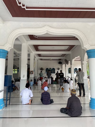 In the city of Trieng Paloh, Indonesia, a group of men are gathered inside a mosque for prayer. The mosque features an arched entryway with white columns, some adorned with blue accents. The ceiling is decorated with wooden panels and white geometric patterns. Inside, men are seated on the floor, facing the front of the mosque, in various stages of prayer. They are dressed in traditional Islamic attire, including sarongs, kufis, and white shirts. Some men are standing, while others are kneeling or bowing. The floor is tiled with white and black lines, reflecting the light from the open space. The environment appears clean and serene, with a few ceiling fans visible and some windows with arched frames in the background.