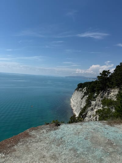The image depicts a cliff overlooking the Black Sea near the village of Dzhankhot, Russia. The cliff face is layered and white, with green pine trees growing on its upper edge and clinging to its slopes. The sea is a deep turquoise color, with gentle ripples on its surface. Several ships are visible in the distance, along with a bridge spanning the horizon. In the foreground, the dry, cracked earth of the cliff top is visible, with patches of light blue discoloration. A small orange object, possibly a buoy or a small boat, floats in the water near the cliff. The sky is a clear blue with scattered white clouds. Two individuals can be seen standing on the cliff edge among the trees.