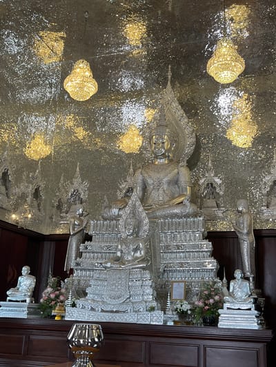 The image shows a silver reclining Buddha statue, possibly located in Muang Pattaya, Thailand, given the cultural context. The Buddha is ornately decorated, with the statue surrounded by smaller silver figures. There are artificial chandeliers on the ceiling. The environment is bright, reflecting the metallic surfaces. Next to the statue there is an object that resembles a vase with flowers. Wooden panels can be seen adjacent to the statue and some silver statues of people kneeling down.