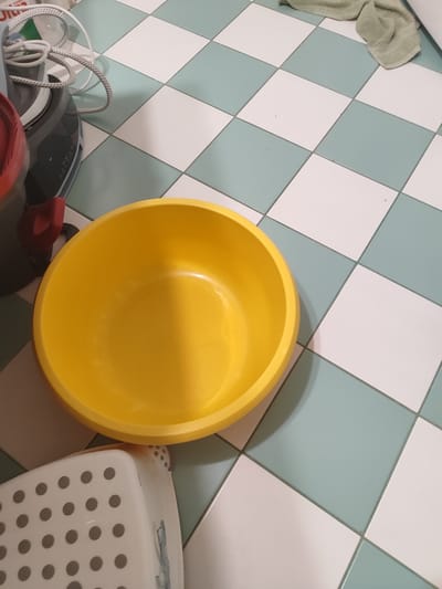 The image shows a yellow plastic basin placed on a checkered floor. The floor tiles are a mix of white and light blue-green. To the left of the basin, there is a portion of a laundry appliance, possibly an iron or a steam station, with a coiled cord visible. A red handle is also attached to this appliance. In the background, a light green towel is draped on the floor. A white plastic stool with dotted perforations is visible in the lower left corner. The lighting suggests an indoor setting, possibly a laundry room or bathroom, during the daytime. No people are present in the image. There is no visible text.