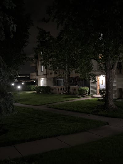 The image shows an exterior view of apartment buildings at night in Vernon Hills, United States of America. The scene is illuminated by pathway lights and the lights from within the apartments, casting a dim glow on the manicured lawns and concrete walkways. Trees with dark foliage are prominent in the foreground and to the sides, partially obscuring the buildings. The apartment buildings themselves are multi-story structures with balconies and visible windows. One doorway is brightly lit, suggesting activity within. The overall atmosphere is quiet and residential.