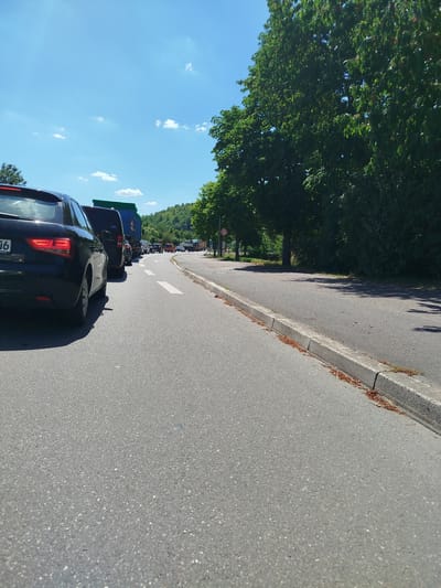The image depicts a traffic jam on a sunny day, likely in the afternoon, given the bright sunlight and shadows. The scene is set on a road bordered by a curb and lush green trees on the right side. The road surface is asphalt, marked with white dashed lines indicating lanes.
In the foreground, the rear of a black Audi car is visible on the left. Further down the road, a line of vehicles, including cars and trucks, are stationary. One truck has a green tarp covering its load. In the distance, a hilly, tree-covered landscape forms the backdrop. On the right side of the road, a circular speed limit sign indicating "20" is visible. The sky is a clear blue with a few scattered white clouds. The overall impression is one of slow-moving traffic on a warm, summer day in a semi-rural or suburban setting. No people are clearly discernible in the image, but the presence of vehicles implies human activity.
Visible text includes "06" on the license plate of the Audi and "169" on a truck. The speed limit sign is also a form of visible text. The location context provided is Lauda-Königshofen, Germany.