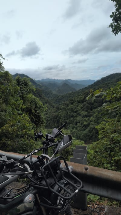 The image is an eye-level shot of a motorcycle parked on the side of a road, with a scenic view of a mountainous, forested landscape in the background. The motorcycle is positioned in the foreground, dominating the lower portion of the frame. It is black and appears to be an adventure or dual-sport model, featuring a prominent front rack, handguards, and mirrors. The details of the motorcycle's front are clearly visible, including the handlebars, headlights, and some of its bodywork, which has a camouflage pattern.
To the right of the motorcycle, a rusty metal guardrail runs along the edge of the road, with a steep drop-off behind it. The guardrail is supported by concrete posts. Beyond the guardrail and the immediate roadside vegetation, the landscape opens up to reveal a vast expanse of rolling mountains covered in dense, lush green forest. The mountains recede into the distance, becoming progressively hazier and bluer due to atmospheric perspective.
The sky above is overcast with a thick layer of gray clouds, suggesting a cloudy or potentially rainy day. The lighting is diffused and muted, contributing to the somewhat somber atmosphere of the scene. The overall impression is one of a remote, natural setting, likely a mountain pass or a road winding through a heavily forested area.
The location is identified as being in Thượng Trạch Commune, Vietnam. This region is known for its mountainous terrain and extensive forests, which aligns with the visual evidence presented in the image. The scene captures a moment of pause during a journey, with the motorcycle serving as a point of reference against the grandeur of the natural environment.