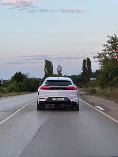 An eye-level, rear-facing, full shot captures a white Lamborghini Urus driving on a paved road at dusk. The car's taillights are illuminated, and its license plate reads "CB MBMB." The road is gray with a white line marking the edge on the left. A dirt shoulder with sparse vegetation is visible to the right, with a small puddle reflecting the sky. In the background, lush green trees and tall, thin trees line the sides of the road. A blue sign with white text in Bulgarian and English points to "Aquapark Sofia" and "Aquapark Sofia." The sky is a soft gradient of light blue and pink with wispy clouds. The overall atmosphere is serene and captures the moment of a drive in the Bulgarian countryside near Ivanyane.