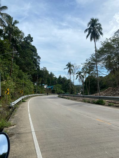 The image depicts a concrete road curving to the left. On the right, a metal guardrail lines the edge of the road. Beyond the guardrail, the terrain slopes downwards, covered in lush green vegetation. Palm trees punctuate the landscape against a blue sky with scattered clouds. The scene suggests a tropical location, possibly near Baan Wang Ta Kien, Thailand, as inferred from the prompt. There are no visible people, actions, or interactions. The environment includes a concrete road, guardrail, vegetation, palm trees, and a cloudy sky.