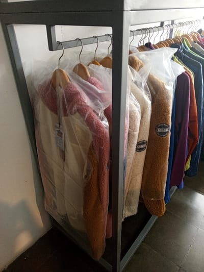 The image captures a segment of a clothing display rack located in Baza, Myanmar (Burma). A dark grey, rectangular metal frame supports a horizontal bar from which multiple garments are suspended on wooden hangers. The visible clothing consists primarily of jackets, many appearing to be fleece or sherpa material, encased individually in clear plastic garment bags. Colors of these jackets include cream, beige, reddish-pink, and various shades of brown. One brown jacket prominently features a circular "KHOOST" brand sticker visible through its plastic covering. Further along the rack, additional garments in various colors such as blue, purple, and green are visible, though less distinctly. The background features a plain white wall, and the floor is composed of dark grey or brown tiles with discernible grout lines. No individuals are present in the scene, and no specific actions or interactions are depicted.