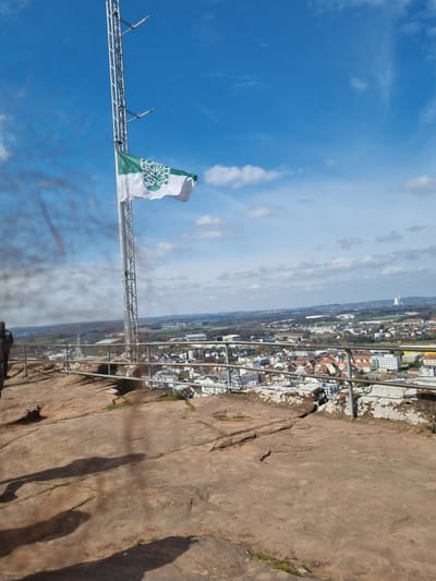 A ground-level, angled-up view shows the city of Homburg, Germany, with a flag waving on a metal platform, a rock formation, and a blue sky. The city, partially obscured by a light haze, is in the middle ground, comprising buildings and a stretch of trees. The foreground features the rock formation, which is light brown and slopes upwards. The metal platform, extending from the edge of the rock, holds a flag of white and green with a leafy emblem. The sky is a deep blue with white clouds and stretches from the left side of the frame upwards, dominating the upper portion of the image. Shadows of the photographer or viewer can be seen on the rock formation.