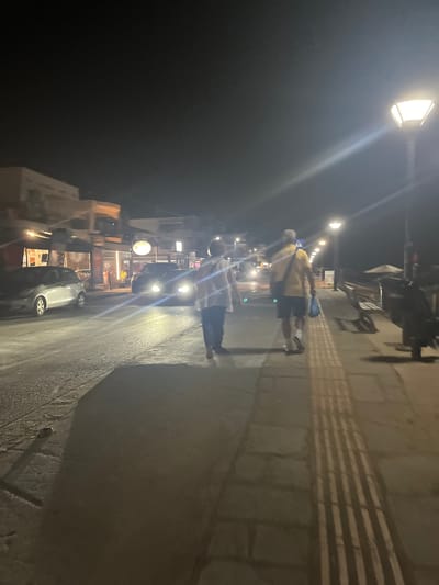 The image depicts a nighttime scene on a street in Orfani, Greece. Two individuals are walking away from the camera on a sidewalk. The person on the left is wearing a light-colored top and dark pants, while the person on the right is wearing a yellow short-sleeved shirt, dark shorts, and carrying a blue bag. They are walking on a paved sidewalk with tactile paving. To the left of the sidewalk is a street with vehicles, including a silver car and other cars with their headlights on. Buildings with illuminated storefronts are visible on the left side of the street. Streetlights illuminate the scene, casting long shadows. In the background, the sky is dark, indicating nighttime.