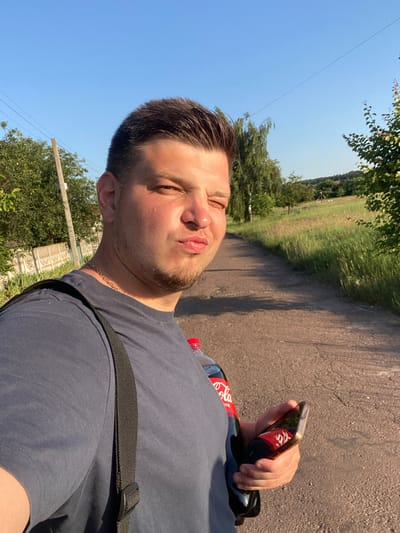 A man is taking a selfie on a dirt road in a rural area, possibly Yablunivka, Ukraine, with a clear blue sky and green trees. He is wearing a dark t-shirt and a gold chain. He is holding a bottle of Coca-Cola and a smartphone in his right hand. The phone screen reflects the Coca-Cola bottle and the man's face. The background shows a grassy field with trees and a gentle hill. The man has short, dark hair and a light beard. His expression is one of playful confidence.